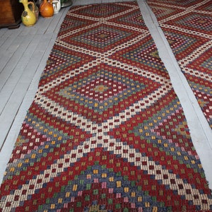 Op de afbeelding: Twee lange, smalle tapijten met een geometrisch patroon in tinten rood, wit, blauw, groen en geel. De tapijten hebben een ruitpatroon met kleinere vierkanten in de ruiten.