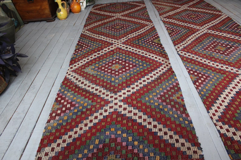 Op de afbeelding: Twee lange, smalle tapijten met een geometrisch patroon in tinten rood, wit, blauw, groen en geel. De tapijten hebben een ruitpatroon met kleinere vierkanten in de ruiten.