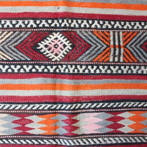 May include: A close-up of a colorful woven rug with a geometric pattern. The rug features stripes of red, orange, gray, and white.