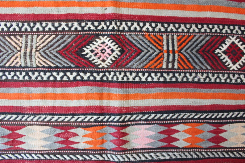 May include: A close-up of a colorful woven rug with a geometric pattern. The rug features stripes of red, orange, gray, and white.
