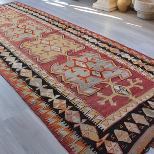 May include: A vintage Turkish rug with a red, orange, yellow, and black geometric pattern. The rug has a fringed edge and is made of wool.