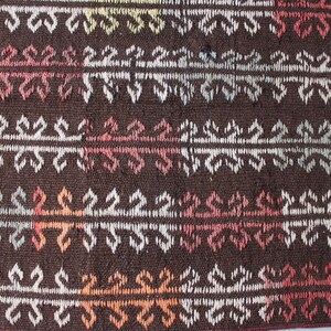 Op de afbeelding: Close-up van een bruin geweven tapijt met een herhalend geometrisch patroon in wit, roze, oranje en blauw.