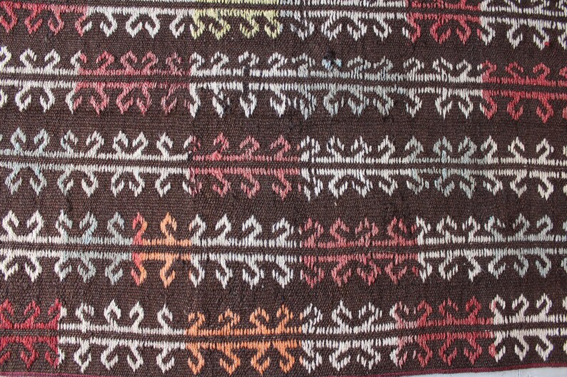 Op de afbeelding: Close-up van een bruin geweven tapijt met een herhalend geometrisch patroon in wit, roze, oranje en blauw.