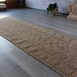 May include: A rectangular, beige-colored runner rug with a detailed geometric pattern. The rug features diamond and floral motifs, with a border design. The rug is placed on a gray wooden floor.