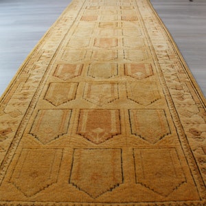 May include: A long, narrow, golden-brown rug with a geometric pattern. The rug has a central field with a repeating pattern of stylized arches, framed by a border with a more intricate design.