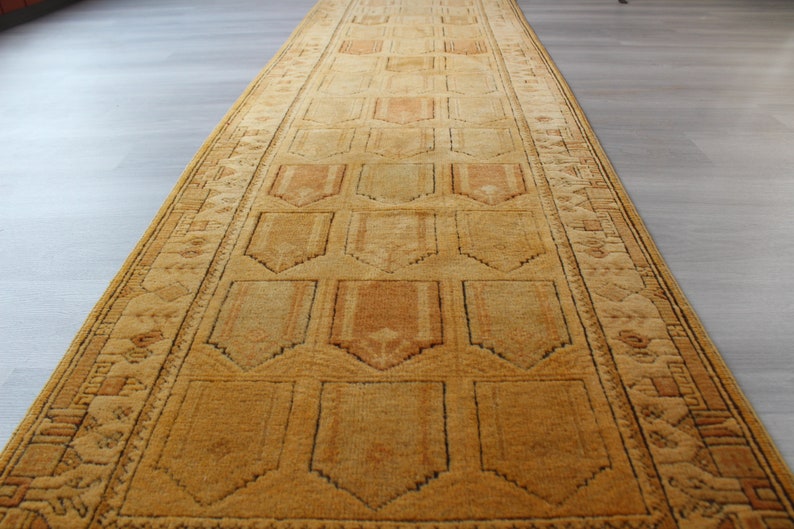 May include: A long, narrow, golden-brown rug with a geometric pattern. The rug has a central field with a repeating pattern of stylized arches, framed by a border with a more intricate design.
