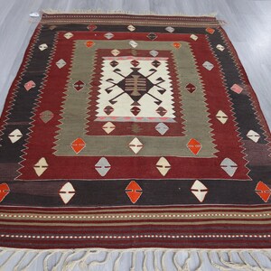 May include: A rectangular, hand-woven rug with a central geometric design in cream, brown, and black. The rug features a border of red, brown, and olive green, with diamond and zig-zag patterns. Cream-colored fringe accents the bottom edge.