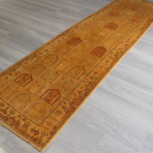 May include: A long, narrow, gold-colored rug with a geometric pattern. The rug has a fringe border and is made of wool.