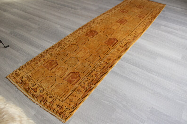 May include: A long, narrow, gold-colored rug with a geometric pattern. The rug has a fringe border and is made of wool.