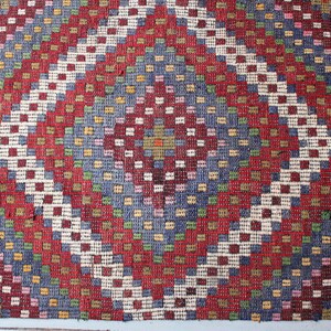 Op de afbeelding: Een close-up van een rood, wit en blauw gepatroneerd tapijt met een geometrisch ontwerp. Het tapijt is gemaakt van geweven stof en heeft een textuur.