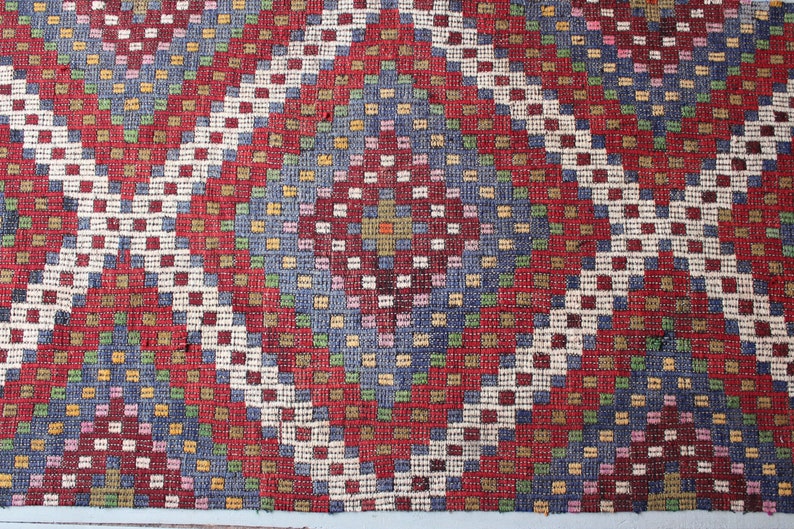 Op de afbeelding: Een close-up van een rood, wit en blauw gepatroneerd tapijt met een geometrisch ontwerp. Het tapijt is gemaakt van geweven stof en heeft een textuur.