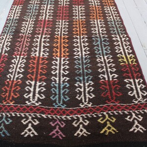 Op de afbeelding: Een lang, smal tapijt met een bruine achtergrond en een kleurrijk geometrisch patroon. Het tapijt heeft een witte rand en franjes aan de uiteinden.