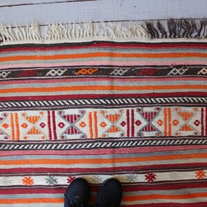 May include: A colorful striped rug with geometric patterns in shades of red, orange, brown, and white. The rug has a fringe border.