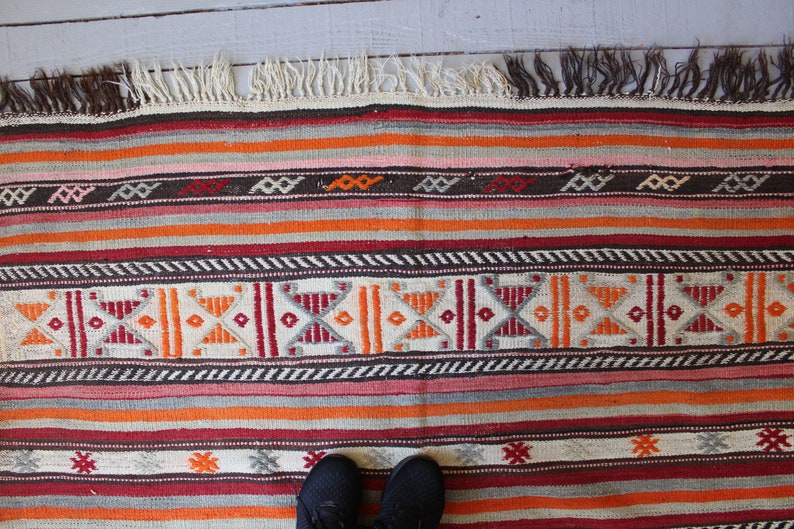 May include: A colorful striped rug with geometric patterns in shades of red, orange, brown, and white. The rug has a fringe border.