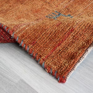 May include: Close-up of a woven rug with a brown, orange, and red color scheme. The rug has a frayed edge and a textured surface.