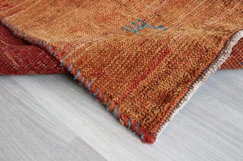 May include: Close-up of a woven rug with a brown, orange, and red color scheme. The rug has a frayed edge and a textured surface.