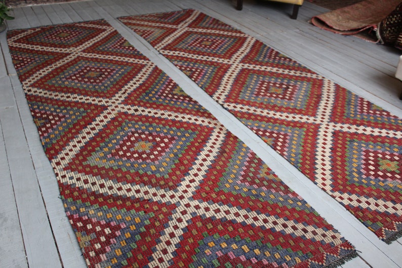 Op de afbeelding: Twee kleurrijke tapijten met een ruitpatroon in tinten rood, blauw, groen en wit. De tapijten zijn gemaakt van geweven stof en hebben een traditionele uitstraling.