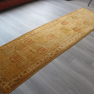 May include: A long, rectangular, gold-colored rug with a geometric pattern. The rug has a border with a repeating design.