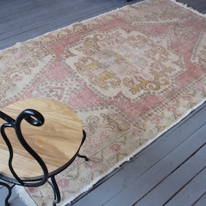 May include: A vintage pink and cream rug with a floral pattern. The rug has a fringed edge and is laid out on a wooden floor.