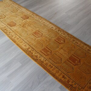 May include: A long, narrow, brown and gold runner rug with a geometric pattern. The rug has a dark brown border with a repeating pattern of small triangles.