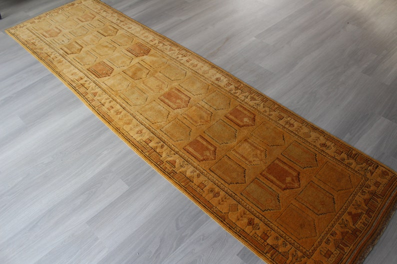 May include: A long, narrow, brown and gold runner rug with a geometric pattern. The rug has a dark brown border with a repeating pattern of small triangles.