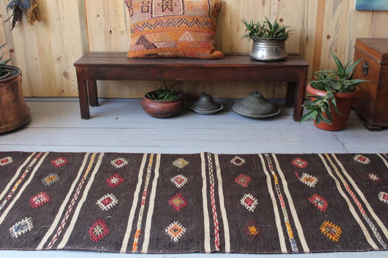 May include: A dark brown woven rug with white stripes and colorful geometric patterns. The rug is laid out on a wooden floor in front of a wooden bench with plants and decorative items.