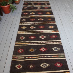 May include: A long, narrow rug with a dark brown background and a geometric pattern in shades of red, blue, yellow, and white. The rug has a traditional, hand-woven look.