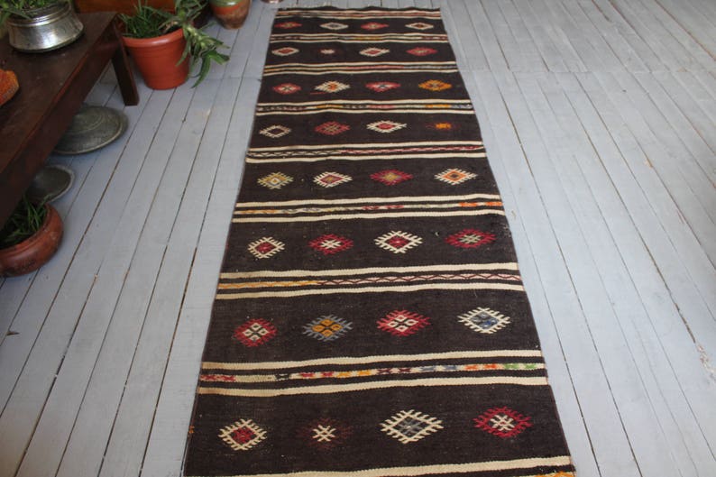 May include: A long, narrow rug with a dark brown background and a geometric pattern in shades of red, blue, yellow, and white. The rug has a traditional, hand-woven look.