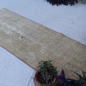 May include: A long, off-white runner rug with a subtle pattern and a red border. The rug is lying on a white surface.