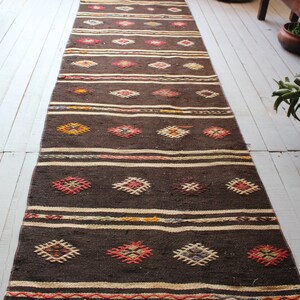 May include: A long, brown runner rug with a geometric pattern in shades of red, orange, yellow, and white. The rug has a woven texture and is made of natural fibers.