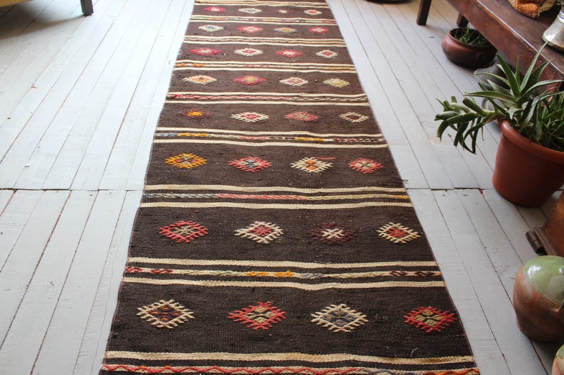 May include: A long, brown runner rug with a geometric pattern in shades of red, orange, yellow, and white. The rug has a woven texture and is made of natural fibers.