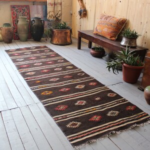May include: A brown and white striped rug with a geometric pattern. The rug is laid out on a white wooden floor in a room with a wooden bench and potted plants.