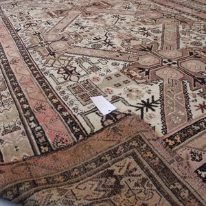 May include: A vintage rug with a cream background and intricate geometric patterns in shades of brown, pink, and black. The rug has a fringed edge and is folded in half.