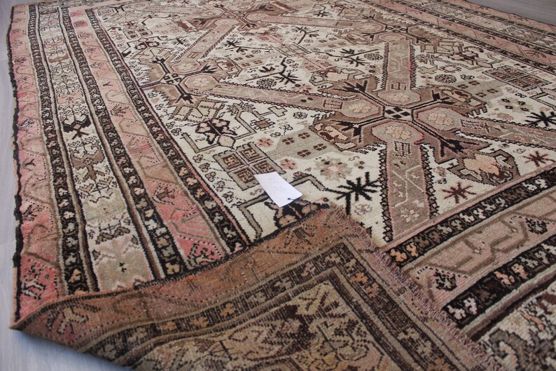 May include: A vintage rug with a cream background and intricate geometric patterns in shades of brown, pink, and black. The rug has a fringed edge and is folded in half.