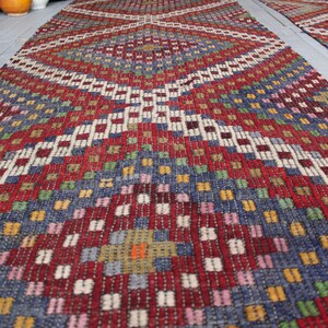 Op de afbeelding: Een kleurrijk geweven tapijt met een geometrisch patroon in tinten rood, blauw, groen, wit en geel. Het tapijt heeft een ruitvormig ontwerp met een centraal vierkant.
