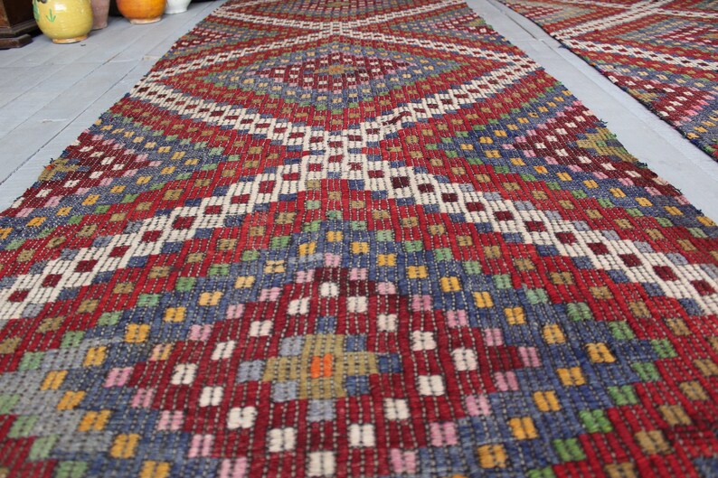 Op de afbeelding: Een kleurrijk geweven tapijt met een geometrisch patroon in tinten rood, blauw, groen, wit en geel. Het tapijt heeft een ruitvormig ontwerp met een centraal vierkant.