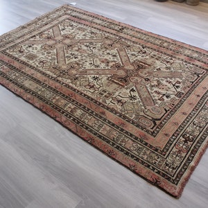 May include: A vintage rug with a cream background and intricate geometric patterns in shades of brown, pink, and black. The rug has a fringed edge and is woven with a traditional design.