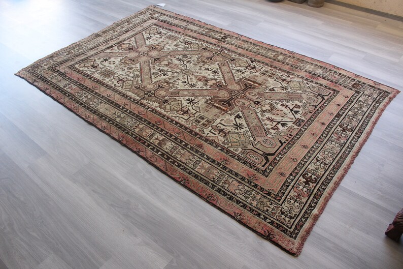 May include: A vintage rug with a cream background and intricate geometric patterns in shades of brown, pink, and black. The rug has a fringed edge and is woven with a traditional design.