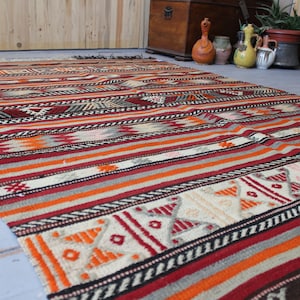 May include: A colorful striped rug with geometric patterns in shades of red, orange, white, and black. The rug is woven with a traditional design and has a fringed edge.