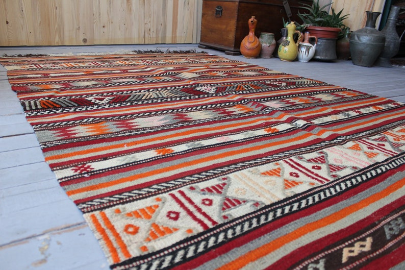 May include: A colorful striped rug with geometric patterns in shades of red, orange, white, and black. The rug is woven with a traditional design and has a fringed edge.
