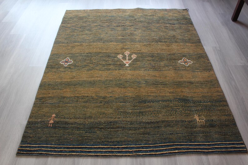 Op de afbeelding: Een groen en bruin vloerkleed met een geometrisch patroon. Het kleed heeft een rand met strepen in blauw, wit en beige.