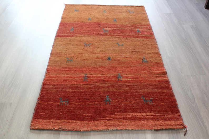 May include: A hand-woven rug with a gradient of orange and red hues. The rug features a repeating pattern of stylized animal figures in a dark blue color.