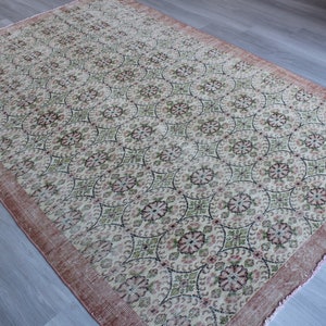 May include: A vintage rug with a cream background and a repeating pattern of pink, green, and black floral motifs. The rug has a pink border with frayed edges.