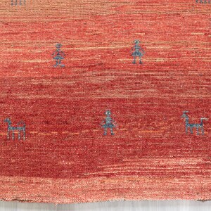 May include: A red and orange handwoven rug with a subtle gradient pattern. The rug features a repeating design of small, stylized figures in a dark blue color.