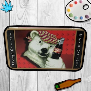 May include: A gold metal tin with a red background and a white polar bear holding a Coca-Cola bottle. The text "Always Coca-Cola" is on the side of the tin.