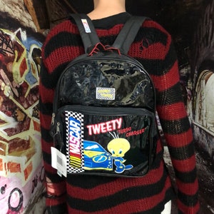 May include: A black and red striped jumper with a black backpack featuring a Tweety Bird design. The backpack has a clear vinyl front panel and a black and white chequered pattern with the word "NASCAR" on it. The backpack also has a yellow Tweety Bird character with the words "Tweety Turbo Charger" on it.