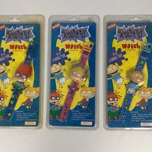 May include: Three Rugrats character watches in packaging. Each watch features a different character: Tommy Pickles, Chuckie Finster, and Angelica Pickles. The watches are blue, pink, and green with a 3-D character on the face.