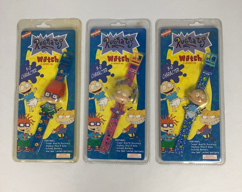 May include: Three Rugrats character watches in packaging. Each watch features a different character: Tommy Pickles, Chuckie Finster, and Angelica Pickles. The watches are blue, pink, and green with a 3-D character on the face.