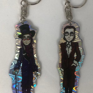 Könnte beinhalten: Zwei holografische Schlüsselanhänger mit Comicfiguren von Männern in Anzügen. Der Schlüsselanhänger links zeigt einen Mann mit Zylinder und Brille, der einen Stock hält. Der Schlüsselanhänger rechts zeigt einen Mann mit langen Haaren und einem dunklen Anzug. Beide Schlüsselanhänger haben einen silbernen Schlüsselring.