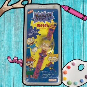 Könnte beinhalten: Eine rosa und gelbe Rugrats-Uhr mit einem 3D-Charakter von Angelica Pickles. Die Uhr verfügt über eine Super-Quarz-Genauigkeit, zeigt Zeit und Datum an, beinhaltet eine Batterie und hat eine einjährige Garantie.